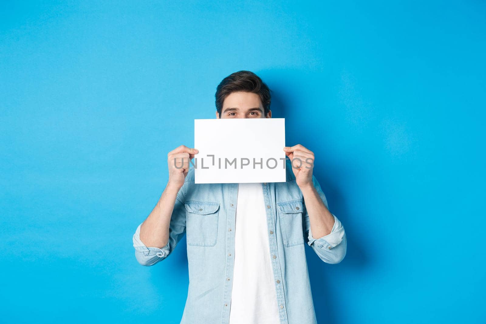 Sneeky handsome guy hiding face behind blank piece of paper for your logo, making announcement or showing promo offer, standing over blue background.