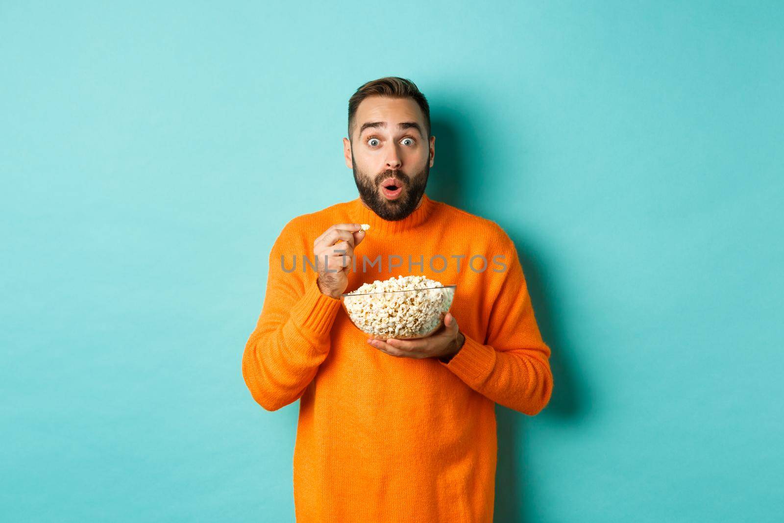 Excited young man watching interesting movie on tv screen, eating popcorn and looking amazed, blue background.