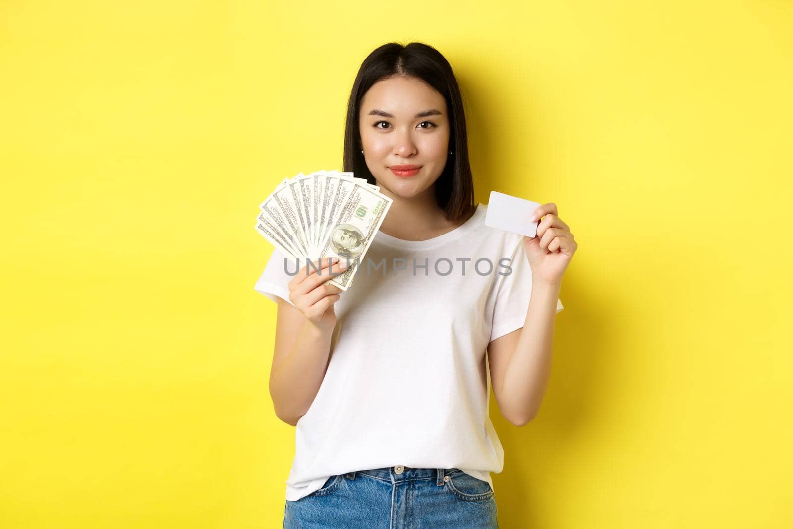 Beautiful asian woman with short dark hair, wearing white t-shirt, showing money in dollars and plastic credit card, standing over yellow background by Benzoix