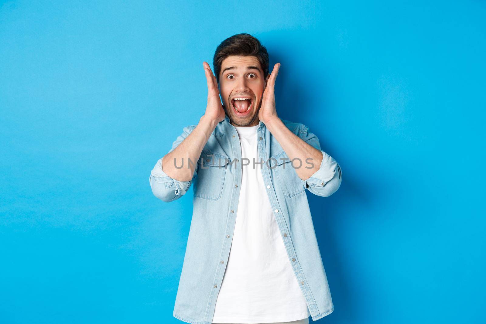 Scared man screaming and looking startled at something, standing against blue background.