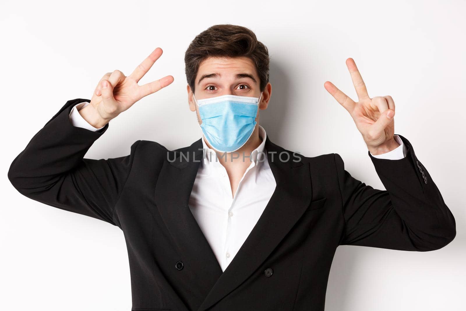Concept of covid-19, business and social distancing. Close-up of happy businessman in suit and medical mask, showing peace signs and smiling, standing against white background by Benzoix