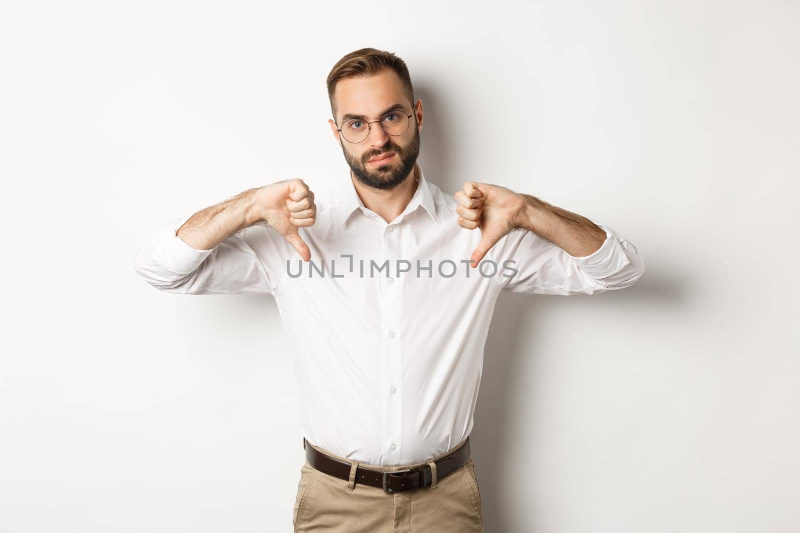 Displeased businessman in glasses showing thumbs down, dislike and disapprove, standing over white background.