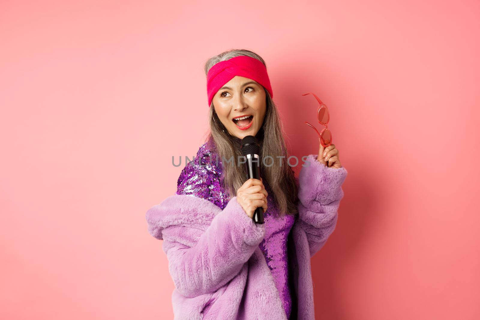Gorgeous asian senior woman singing karaoke in mic, perform song and looking happy, standing over pink background by Benzoix