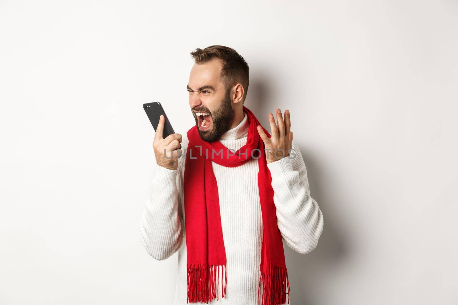 Angry man shouting at smartphone with mad face, standing furious against white background.