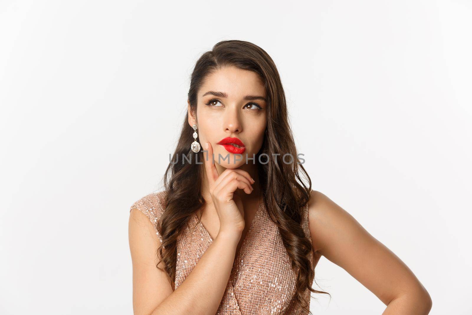 Concept of New Year celebration and winter holidays. Close-up of elegant woman with red lips and dress, looking at upper left corner and thinking, standing over white background.