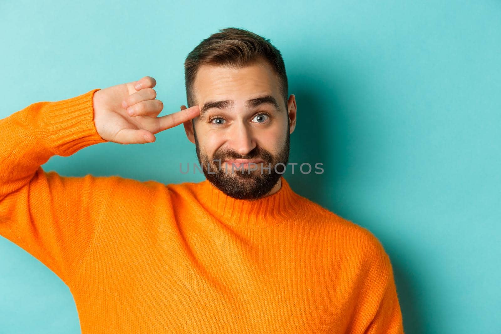 Close-up of man expressing disdain, scolding person, pointing finger at head and looking at camera, standing over light blue background.