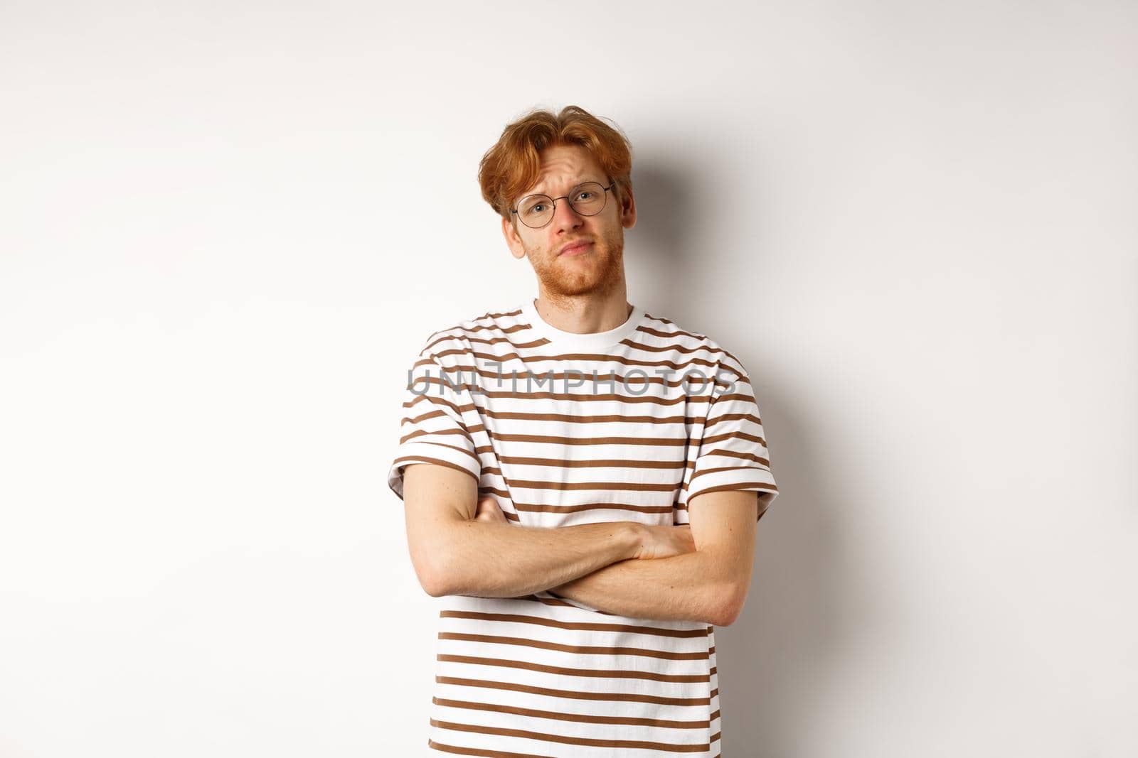 Teenage redhead guy in glasses cross arms on chest, looking skeptical and unamused at camera, standing over white background.