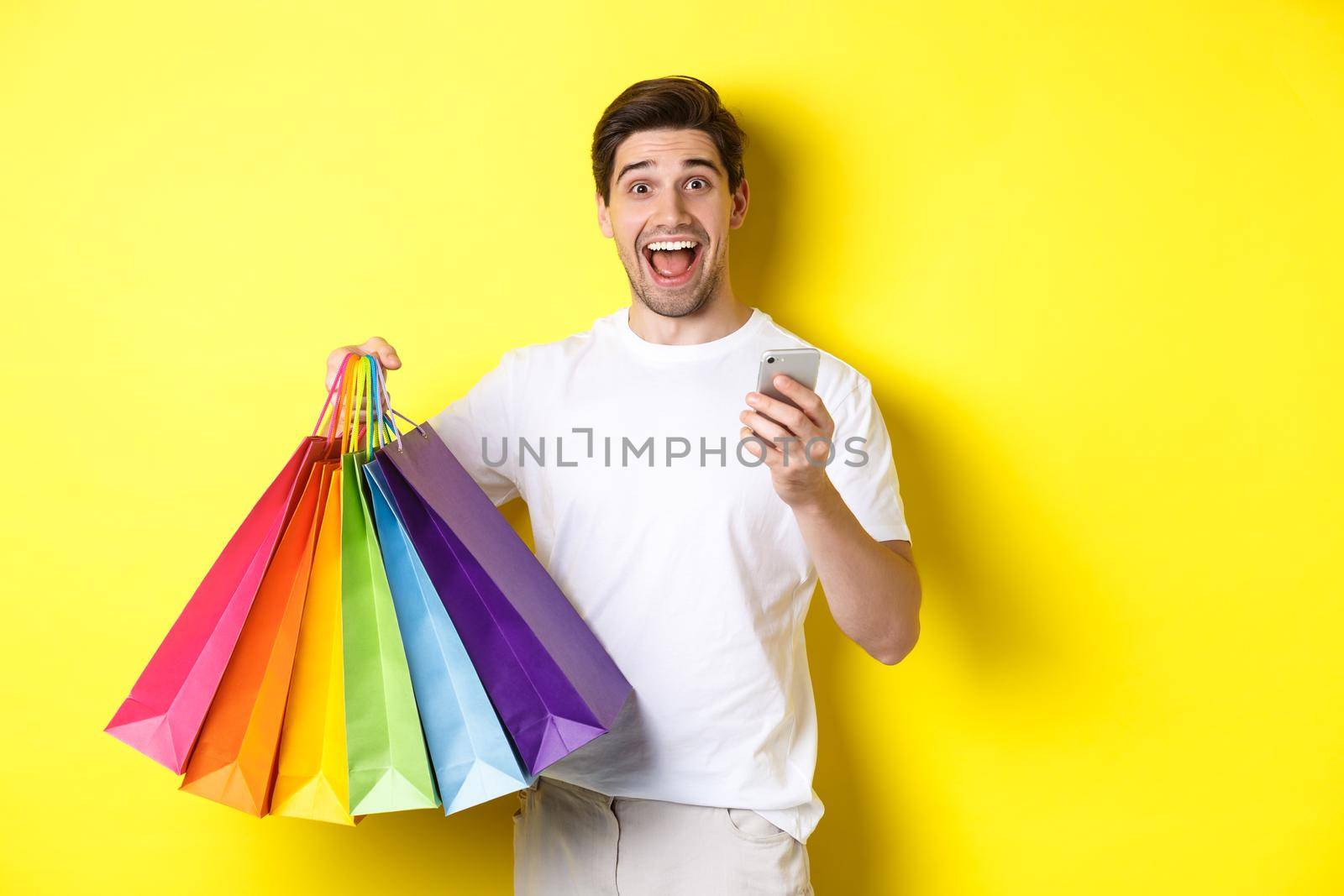 Image of happy man receive cashback for purchase, holding smartphone and shopping bags, smiling excited, standing over yellow background.