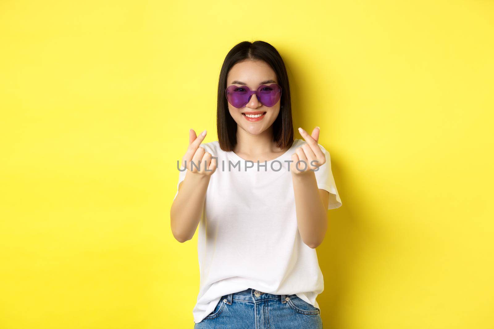 Fashion and lifestyle concept. Attractive asian woman in heart-shape sunglasses, showing finger hearts and smiling happy at camera, standing over yellow background.