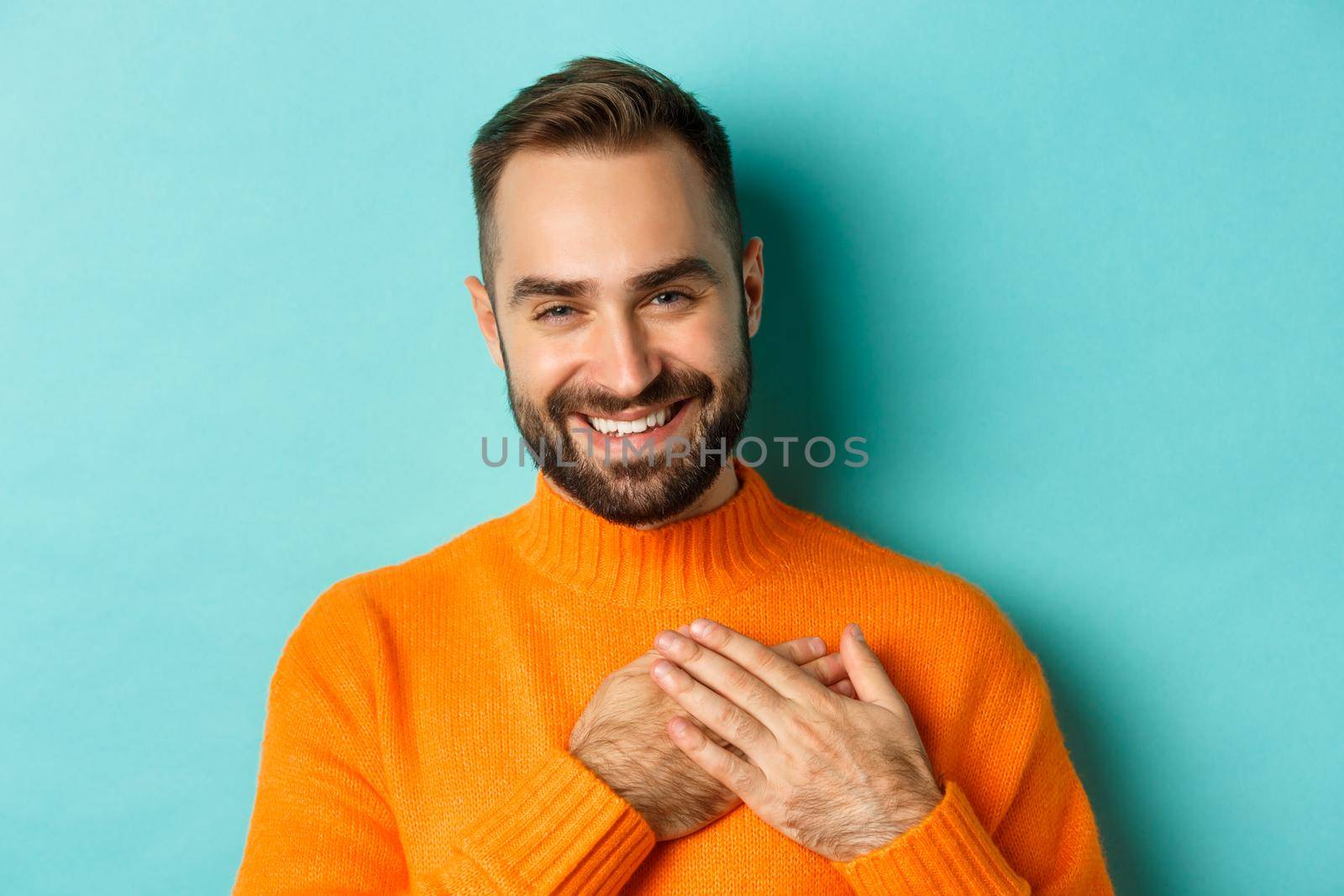 Close-up of handsome young man saying thank you, holding hands on heart and smiling grateful, standing over light blue background by Benzoix