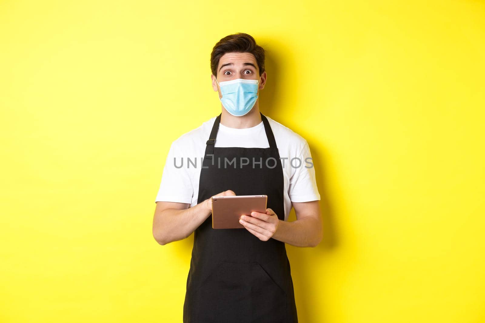 Concept of covid-19, small business and pandemic. Waiter in black apron and medical mask taking order, holding digital tablet, standing over yellow background.