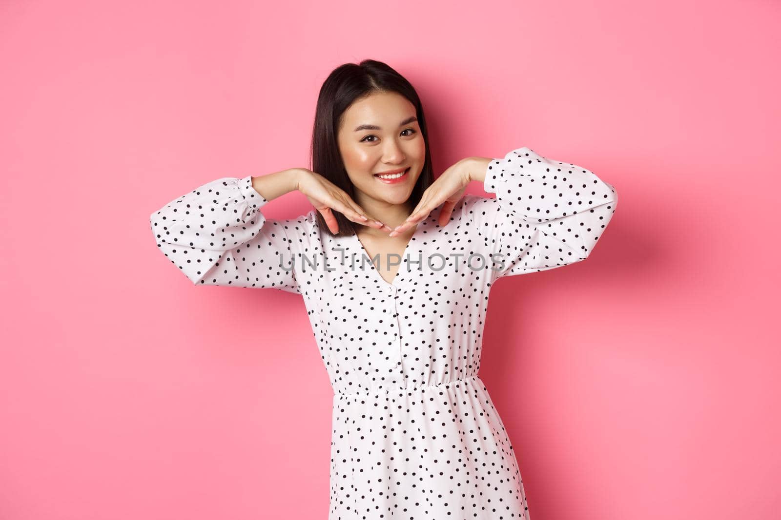 Lovely asian woman showing her beautiful clean face, smiling pretty, standing in dress against pink romantic background.