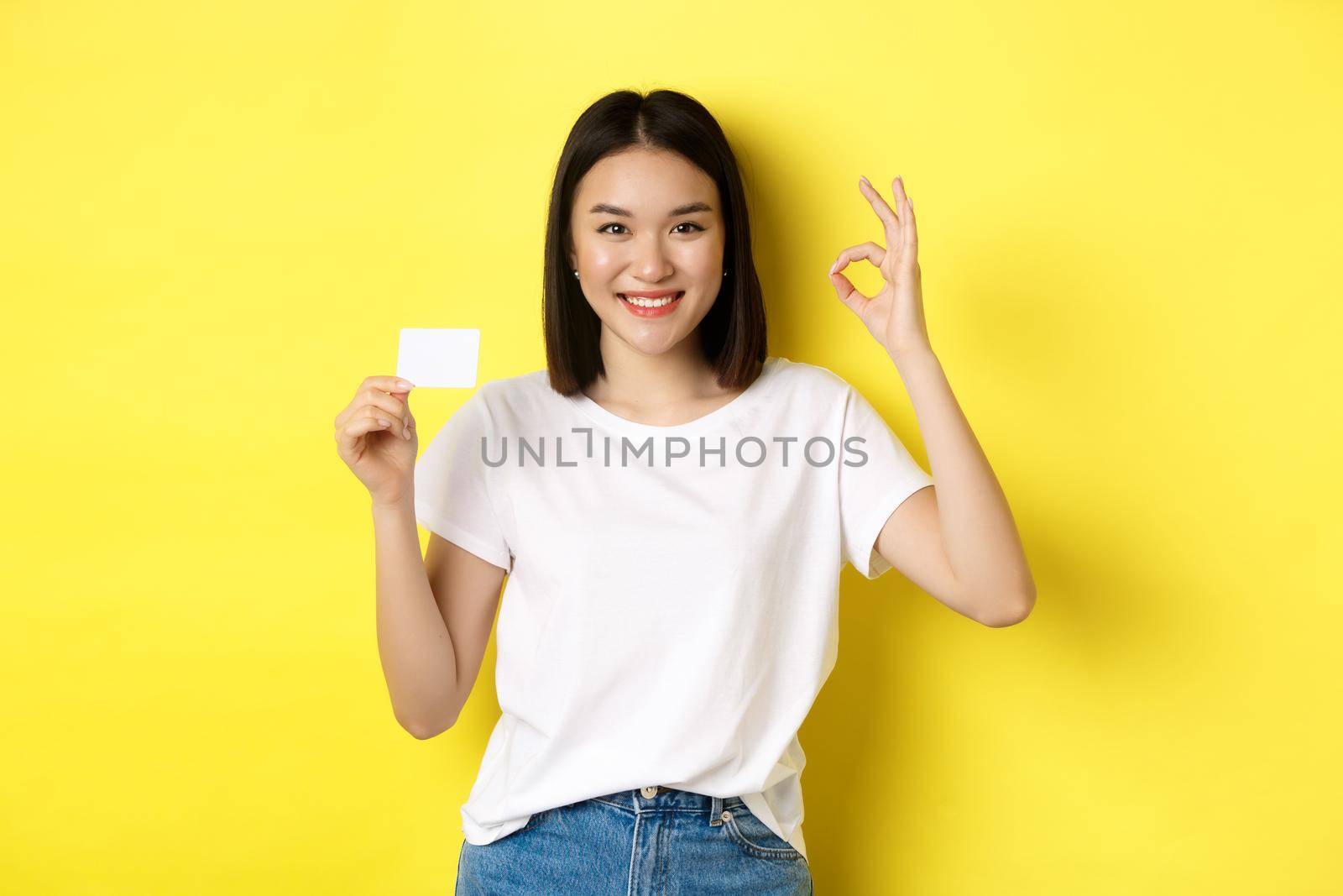 Young asian woman in casual white t-shirt showing plastic credit card and okay gesture, recommend bank, smiling at camera, yellow background by Benzoix