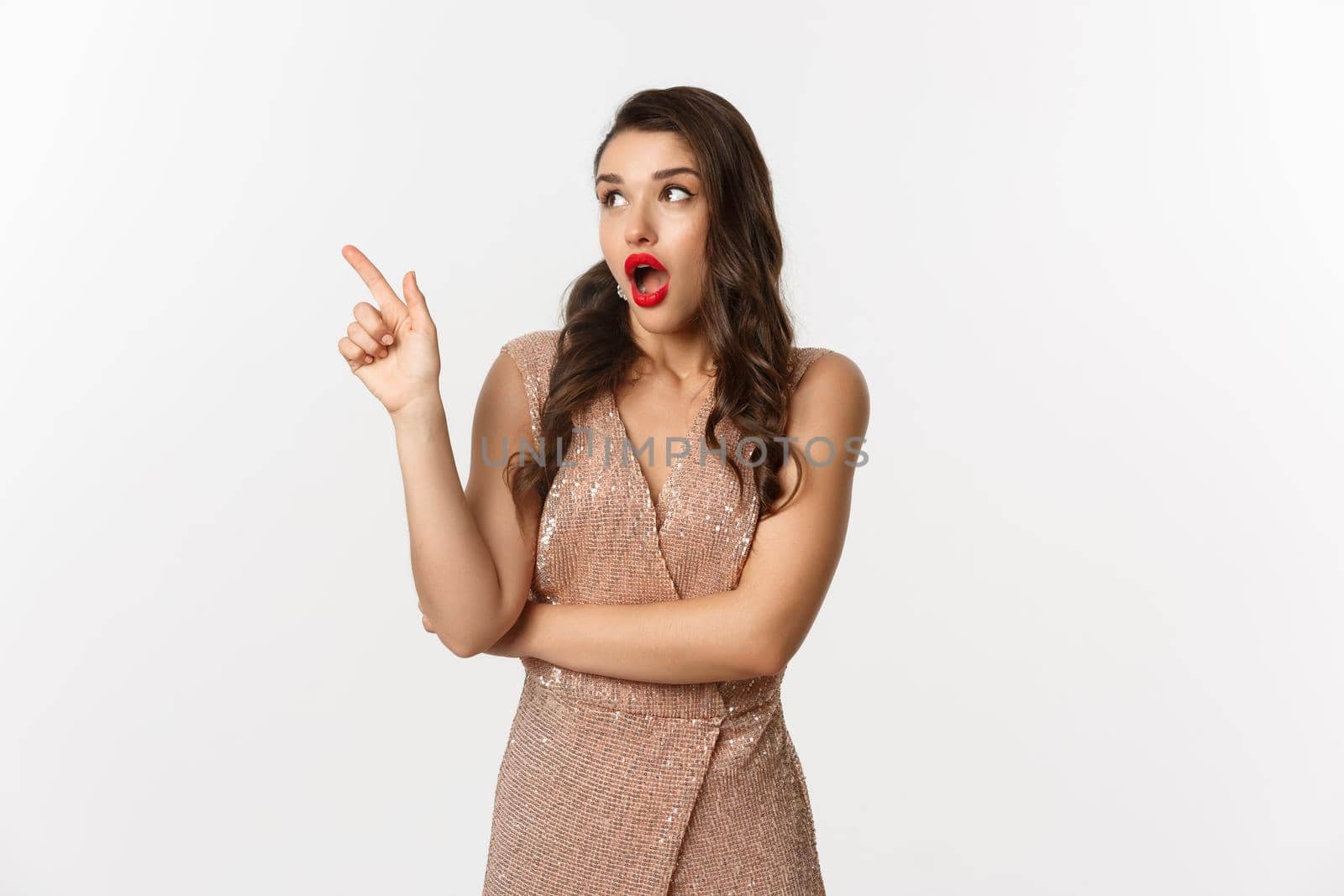 Christmas, holidays and celebration concept. Amazed fashionable woman in party dress and red lipstick, pointing and looking left at promo offer, standing against white background by Benzoix