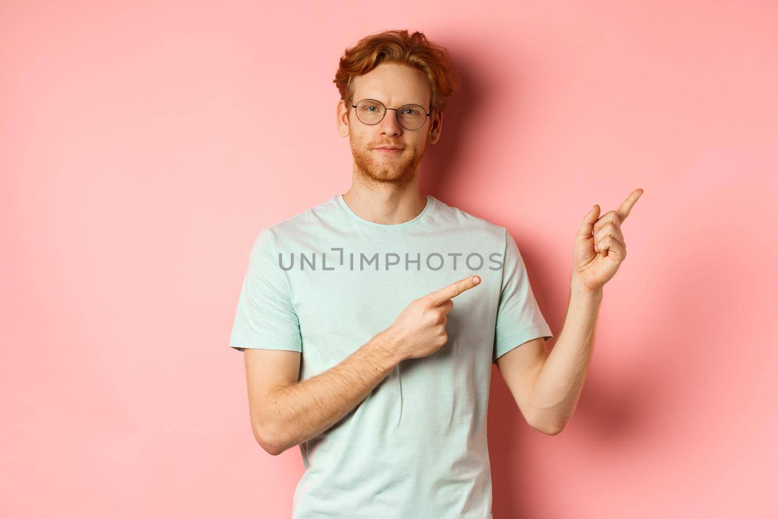 Confident young man with ginger hair and beard, smiling pleased and pointing fingers at upper right corner, invite or demonstrate something, standing over pink background.