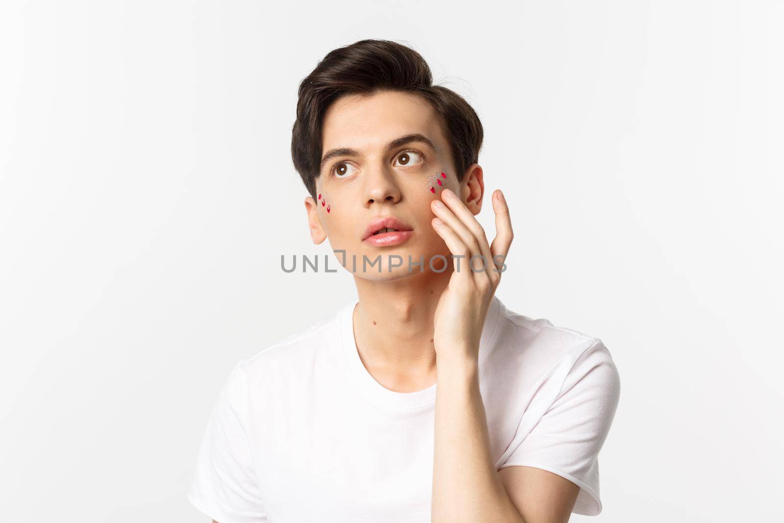People, lgbtq community and lifestyle concept. Beautiful young gay man applying glitter under eyes for pride party, standing over white background.