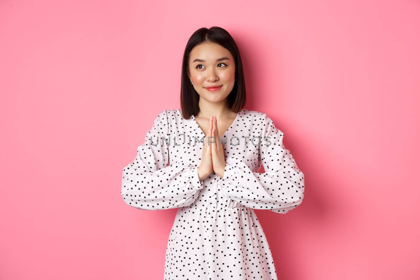 Beautiful angelic asian woman smiling, holding hands in pray and looking left at copy space with innocent cute gaze, standing over pink background.