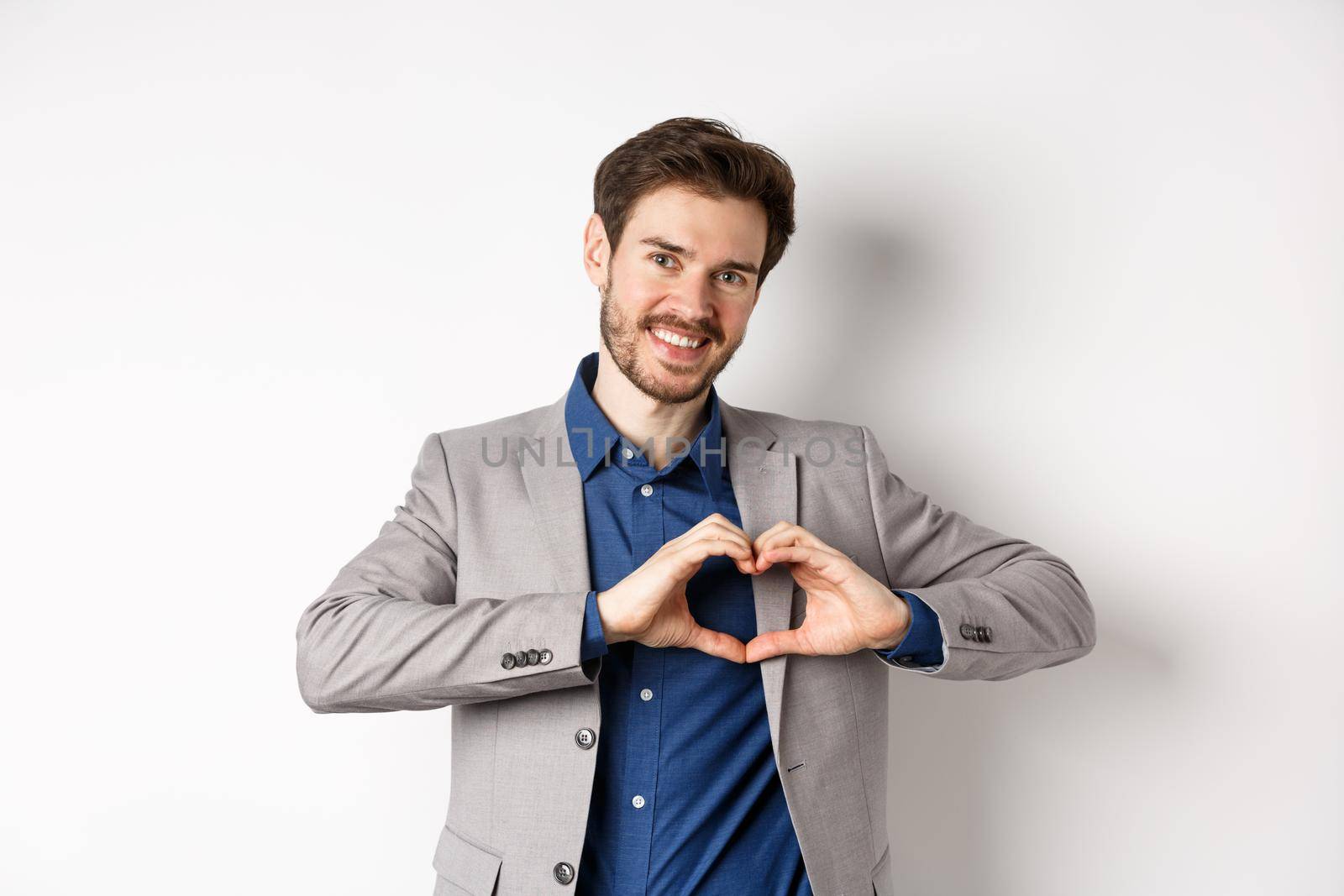 Romantic man in suit showing heart sign and smiling, love his girlfriend, standing on white background, making confession to lover by Benzoix