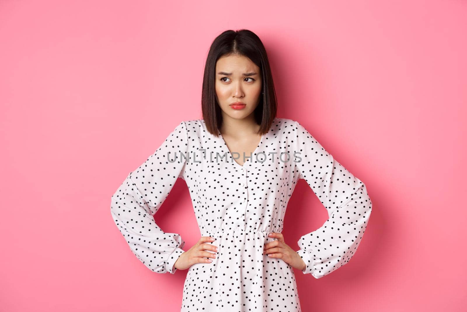 Sad and concerned asian woman having problem, holding hands on waist and looking left with upset face, standing in dress against pink background by Benzoix