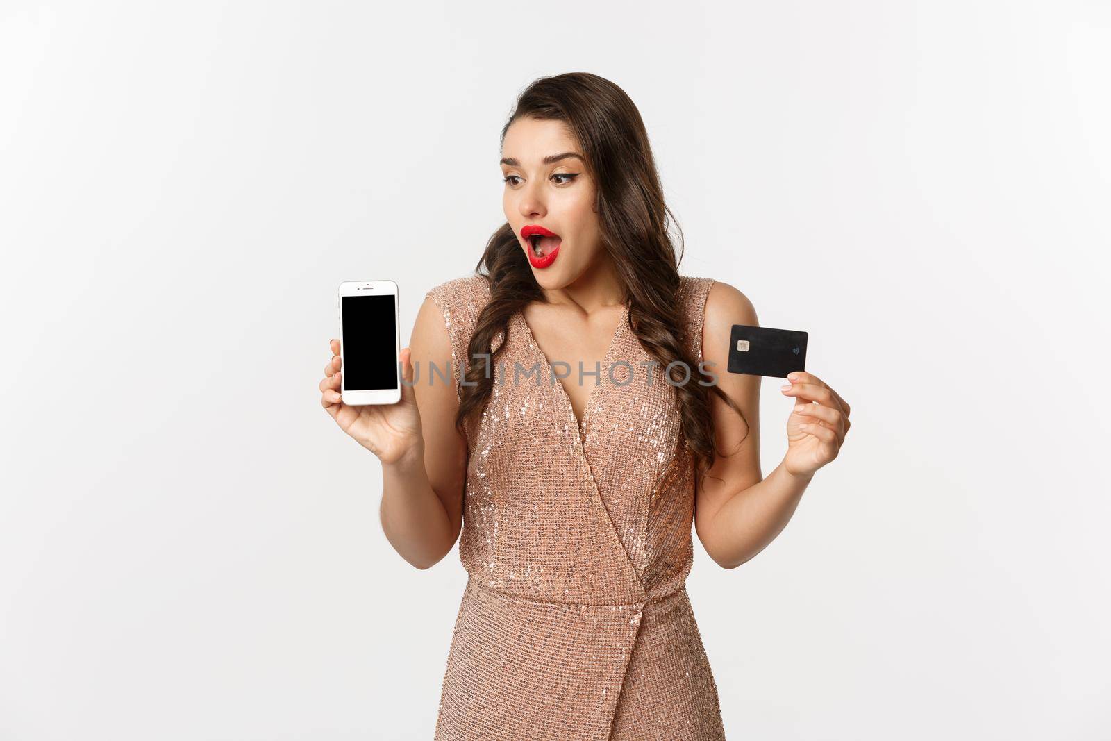 Online shopping and holidays concept. Elegant woman in party dress looking amazed at phone screen, showing internet store and credit card, white background.