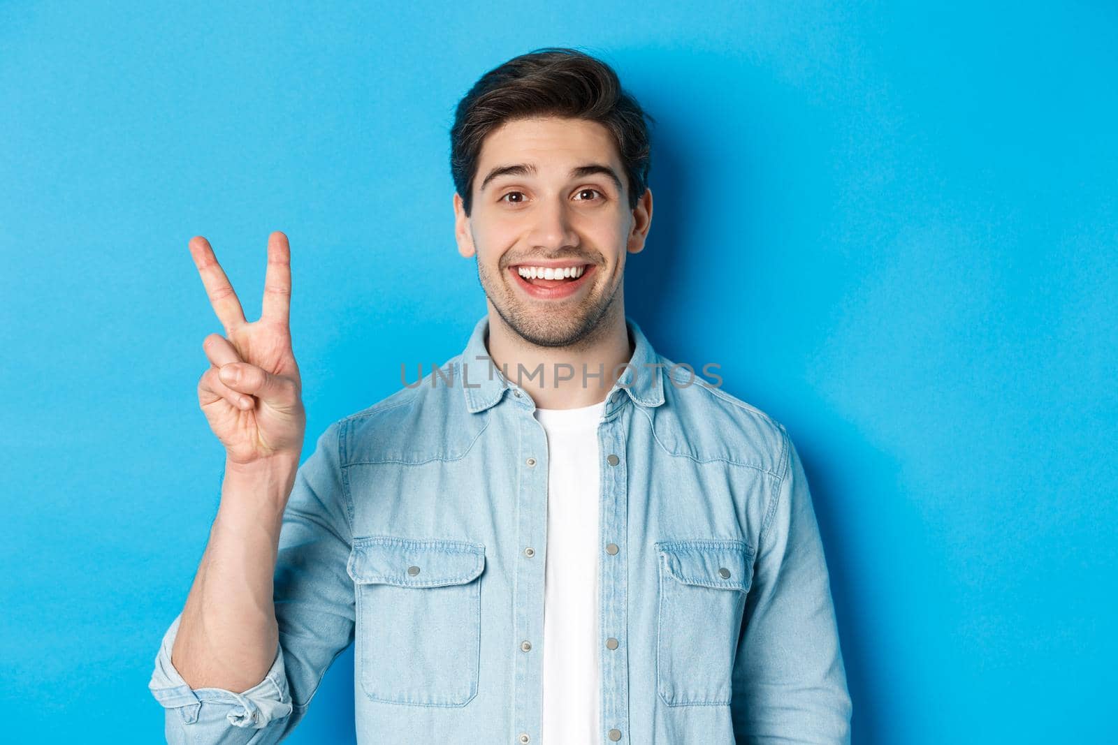 Close-up of handsome man smiling, showing fingers number two, standing over blue background.