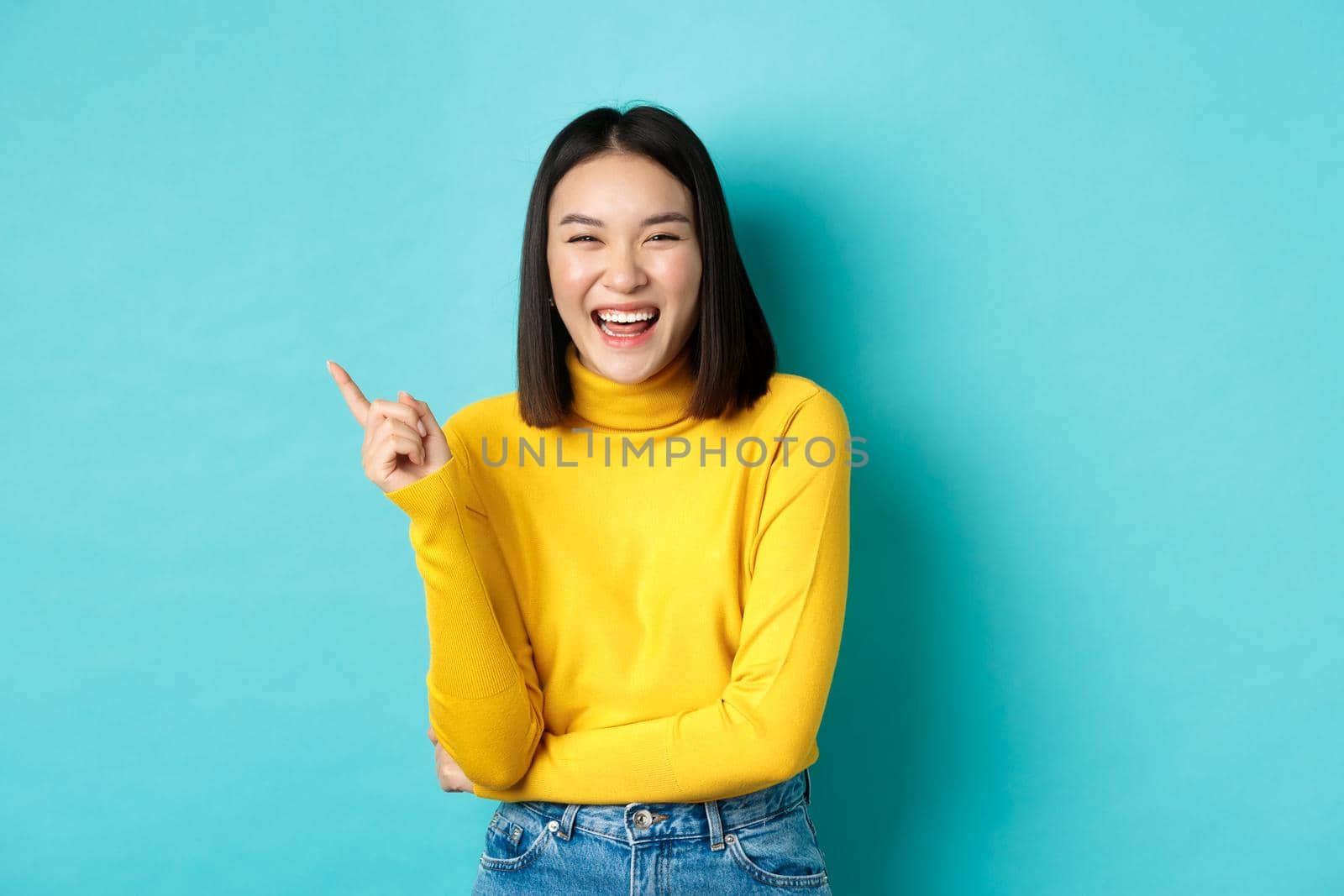 Image of carefree pretty korean girl laughing and looking happy, pointing finger at upper left corner promo, standing against blue background by Benzoix