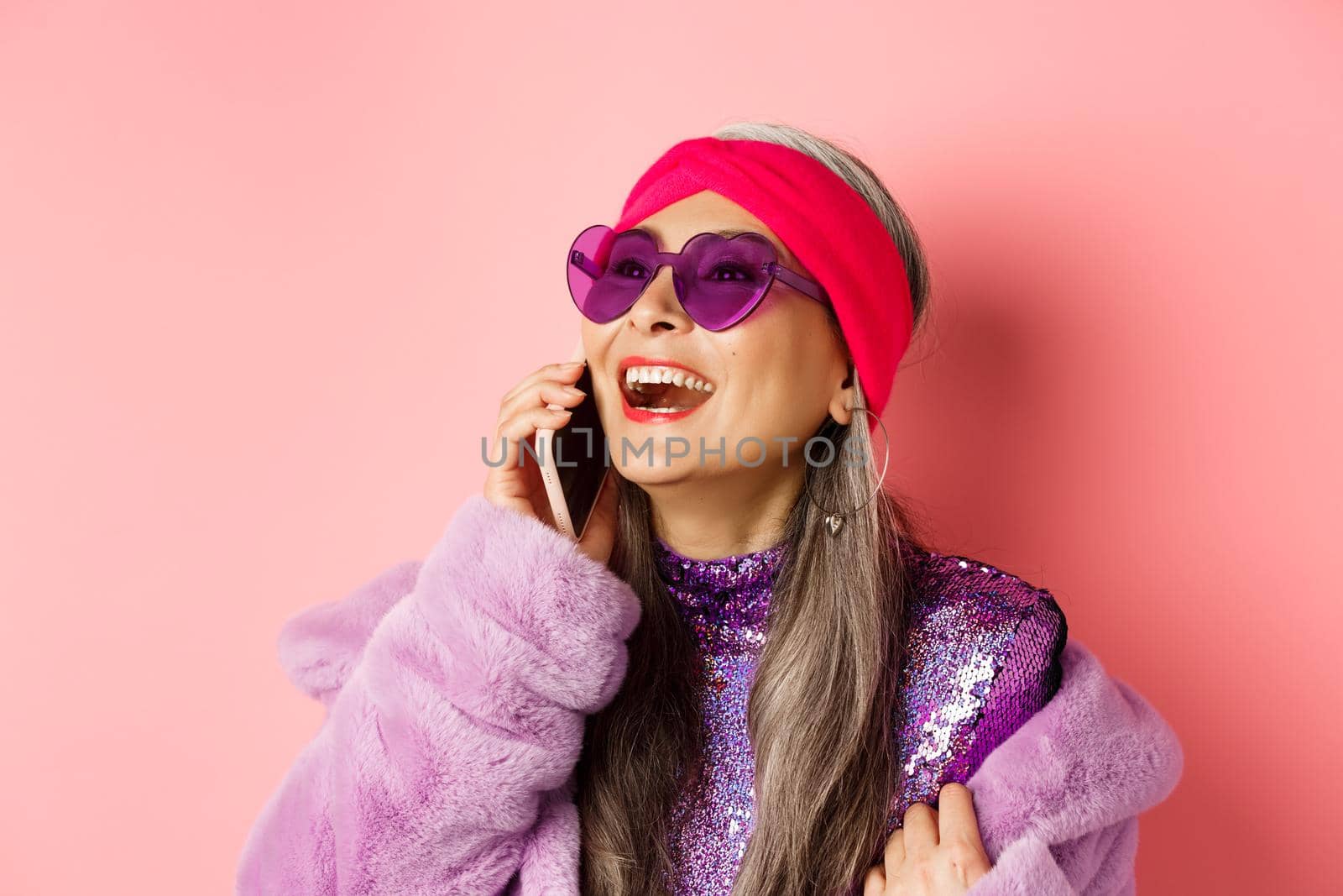 Fashion. Beautiful asian senior woman in trendy sunglasses and headband laughing, talking on mobile phone and looking aside, pink background by Benzoix