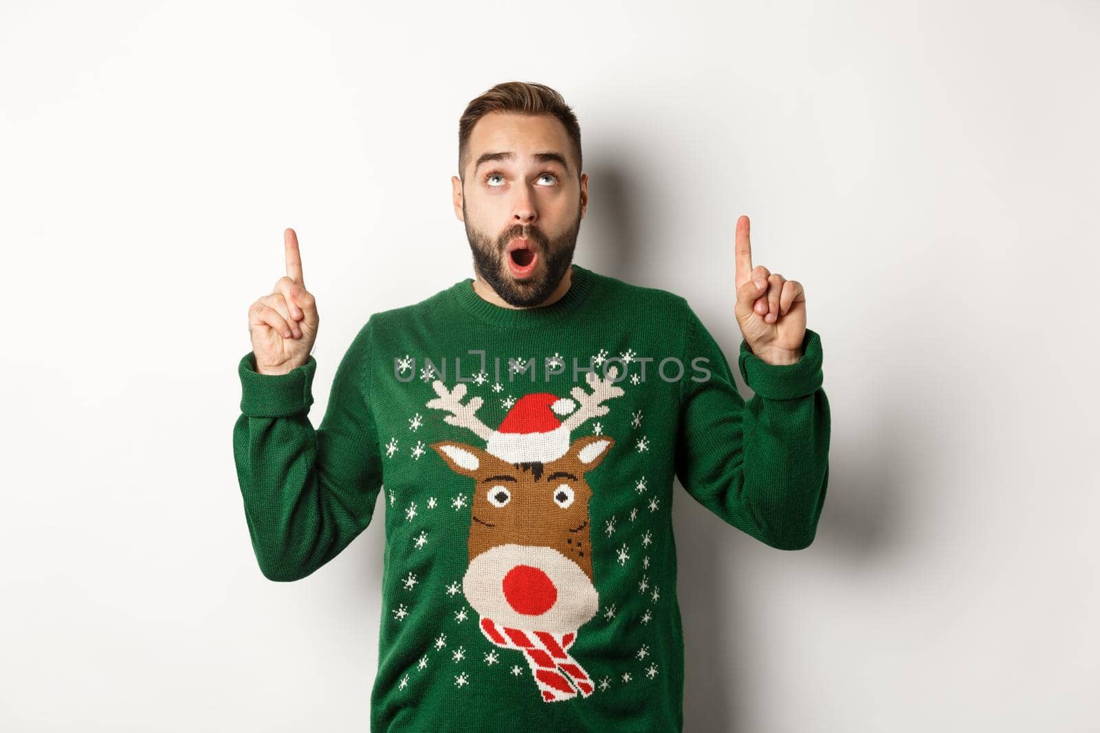 Winter holidays and christmas. Amazed handsome man in new year sweater pointing fingers up, showing logo, standing over white background. Copy space