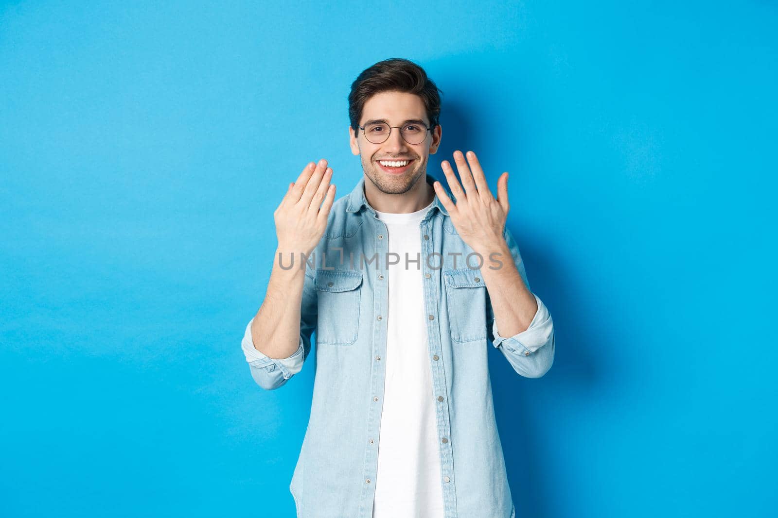 Handsome young man in glasses showing manicure hands and smiling, taking care of nails, standing over blue background by Benzoix