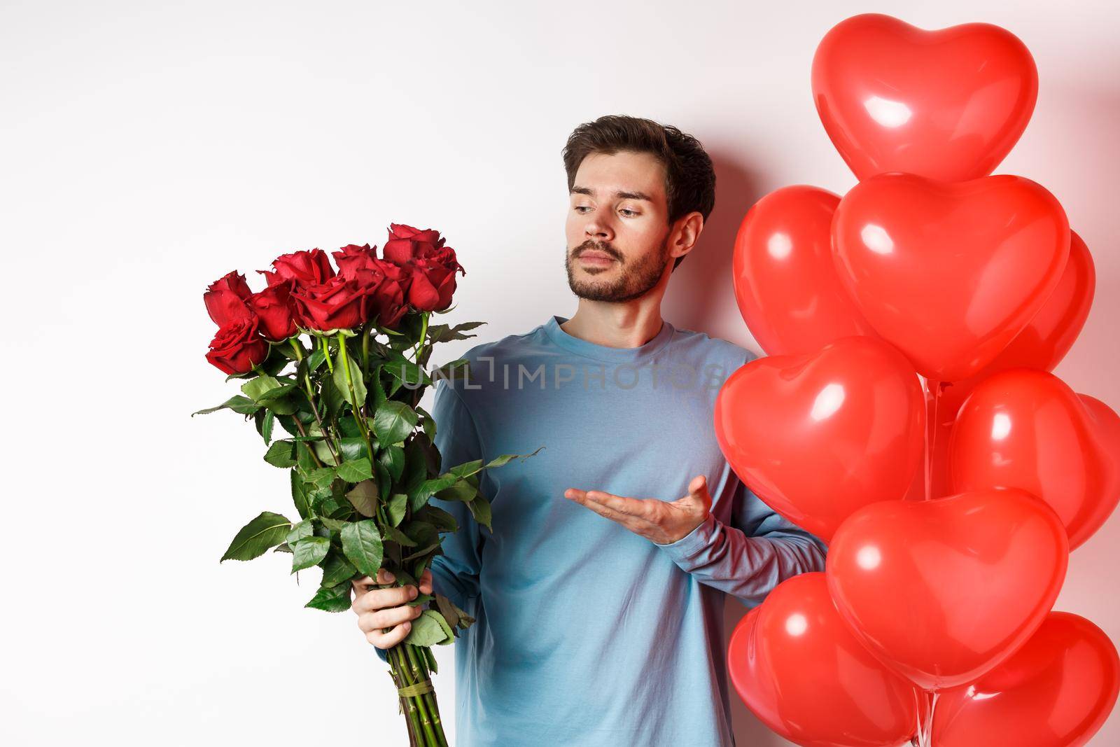 Romantic guy showing bouquet of red roses, pointing at flowers and standing near hearts balloons on Valentines day, prepare gifts for his lover, white background.