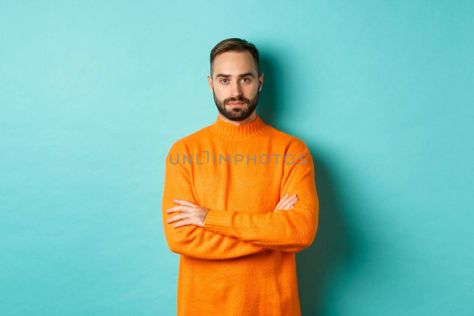 Confident young man looking determined, cross arms on chest, wearing orange winter sweater, standing against turquoise studio background by Benzoix