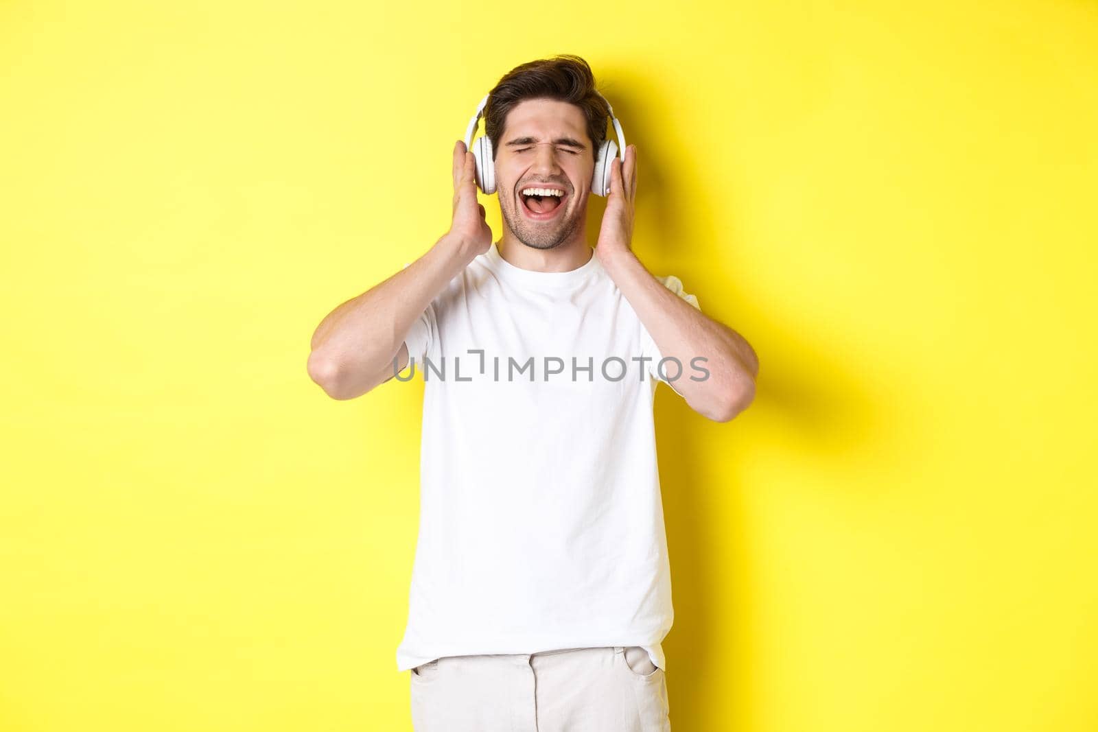 Happy guy listening music in new headphones, buying earphones on black friday, standing over yellow background.