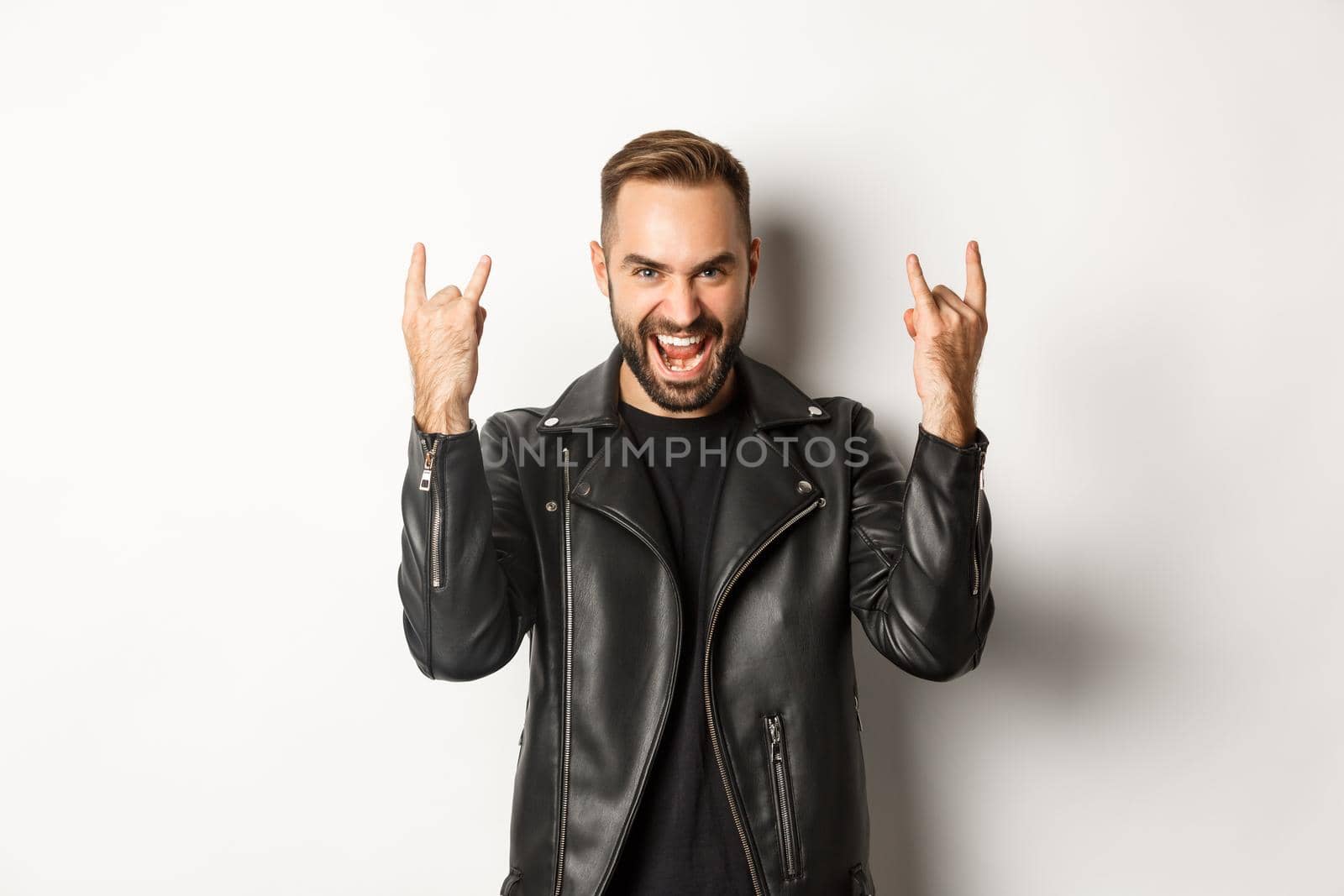 Cool adult man in black leather jacket, showing rock on gesture and tongue, enjoying music festival, standing over white background by Benzoix