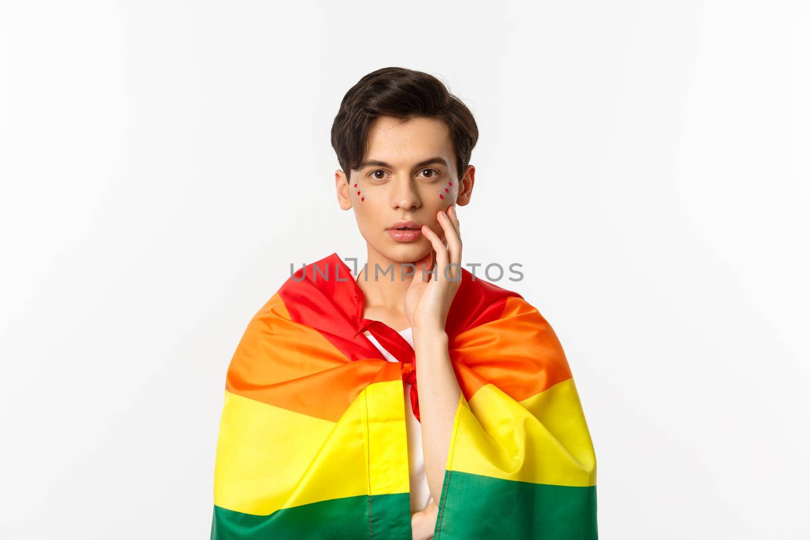 Beautiful young gay man wearing lgbt pride flag and glitter on face, touching cheek and gazing sensual at camera, white background.