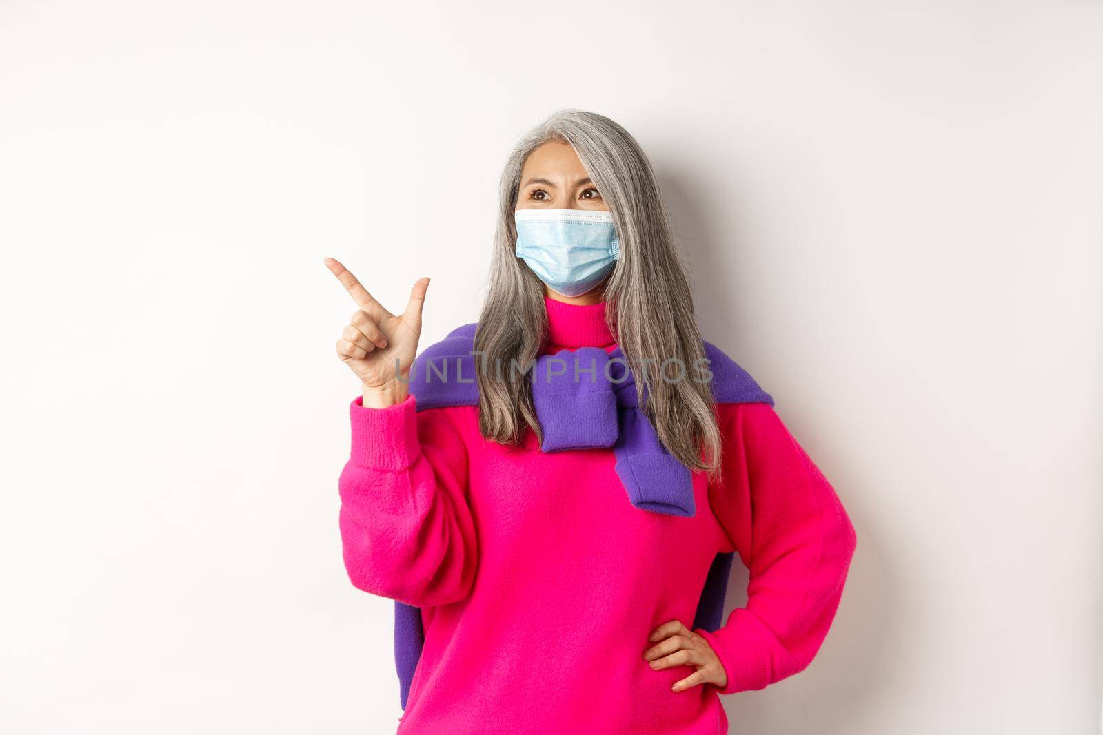 Covid-19, quarantine and lockdown concept. Stylish asian grandmother in face mask pointing and looking at upper left corner, showing logo and smiling, white background by Benzoix