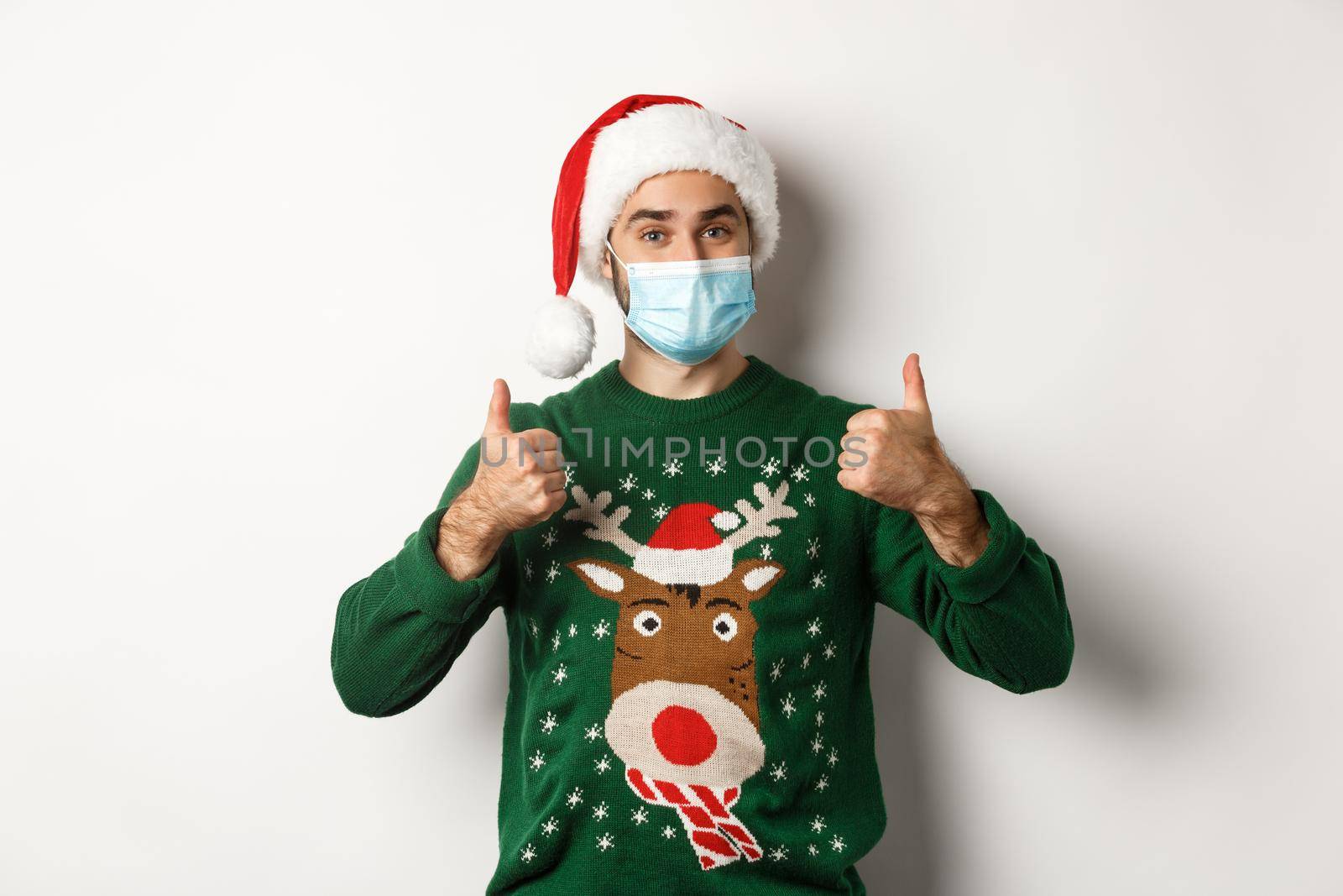 Christmas during pandemic, covid-19 concept. Satisfied man in Santa hat and medical mask showing thumb up in approval, praising something, standing over white background.