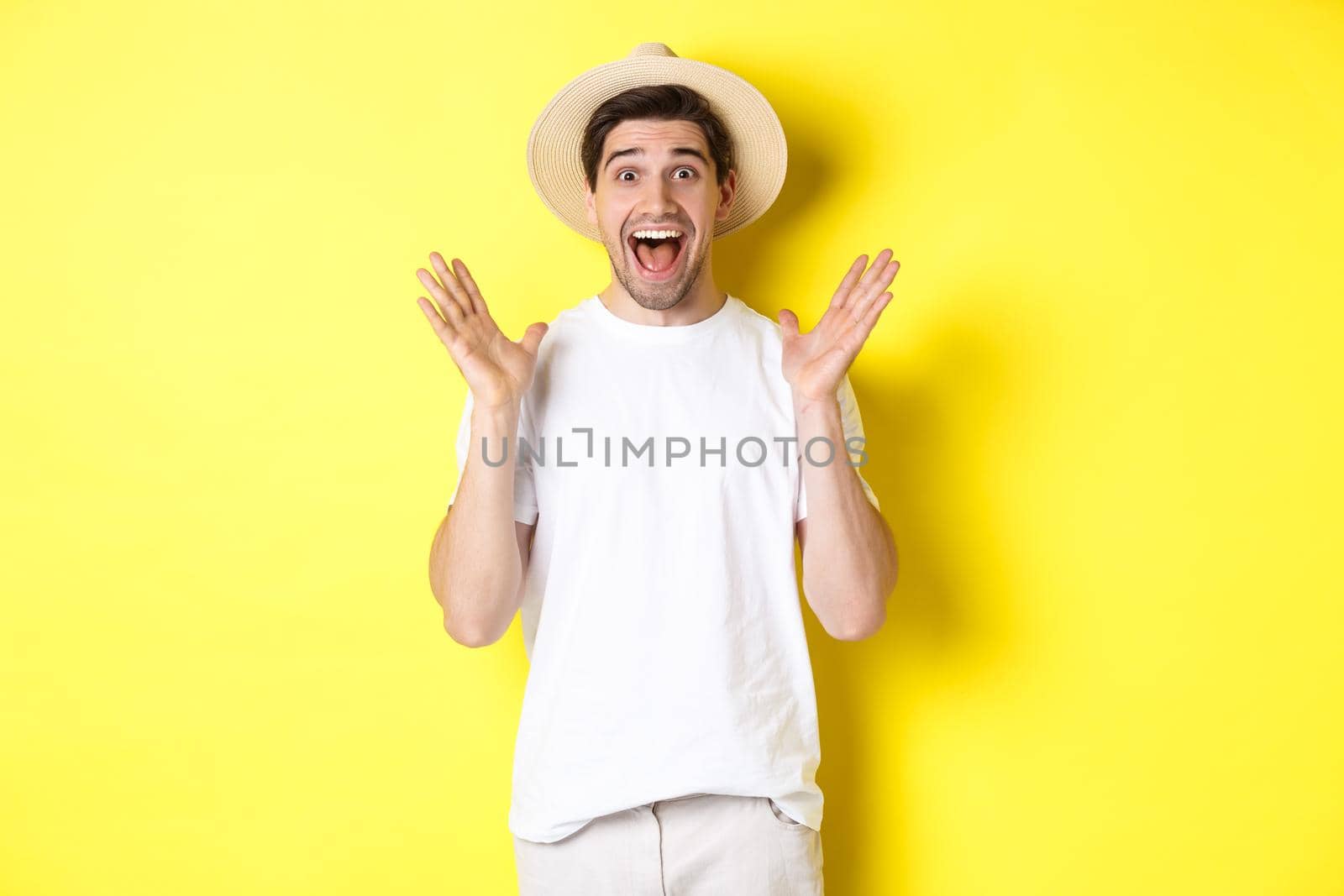Concept of tourism and summer. Happy young man in straw hat looking amazed, reacting to surprise, standing over yellow background.