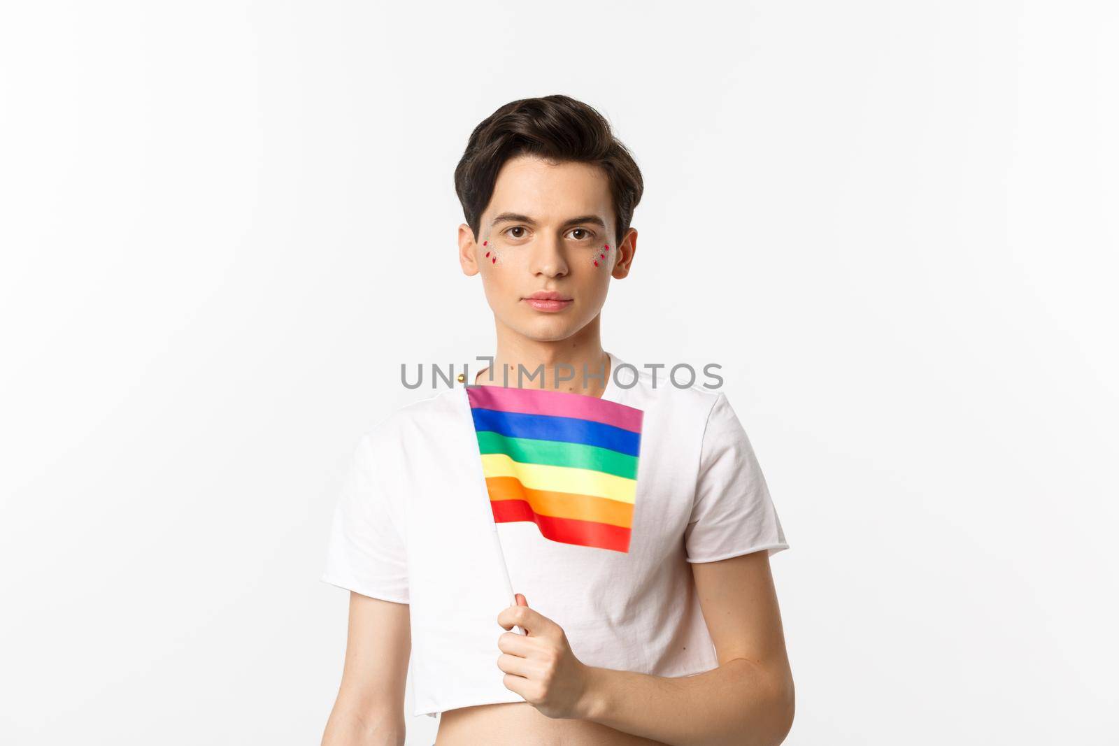 Pride and lgbtq concept. Waist up shot of attractive anrogynous man holding rainbow flag, having glitter on face and looking at camera, standing over white background.