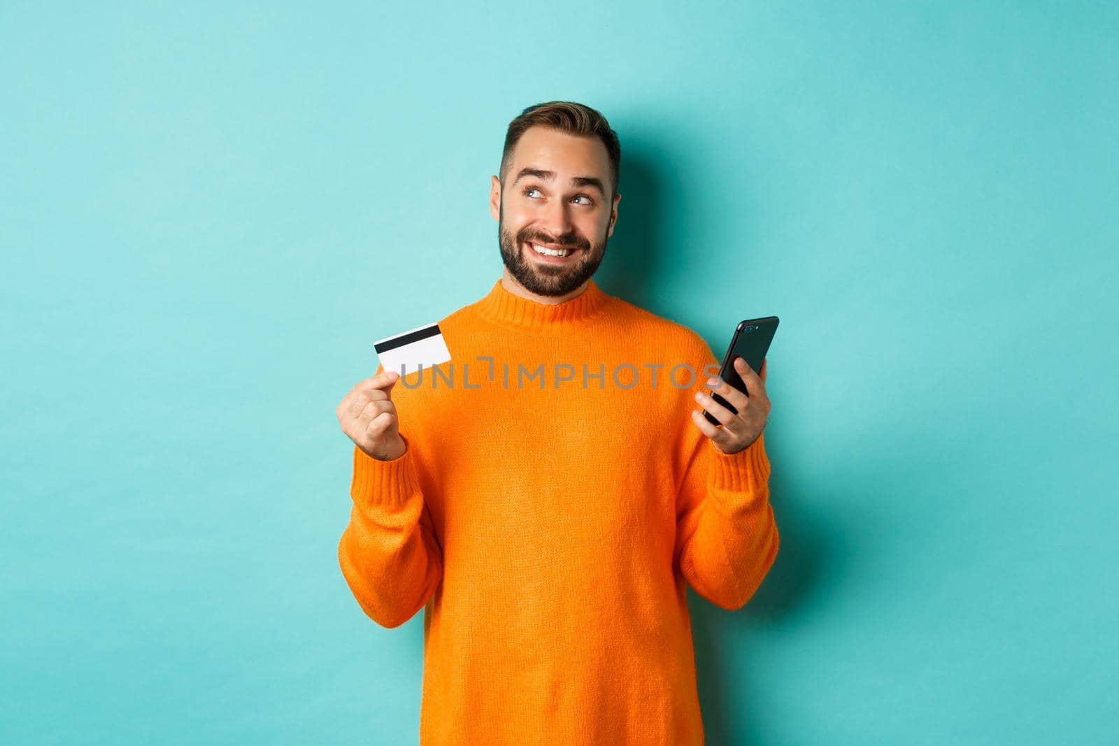 Online shopping. Handsome man thinking, holding smartphone with credit card, paying in internet store, standing over light blue background by Benzoix