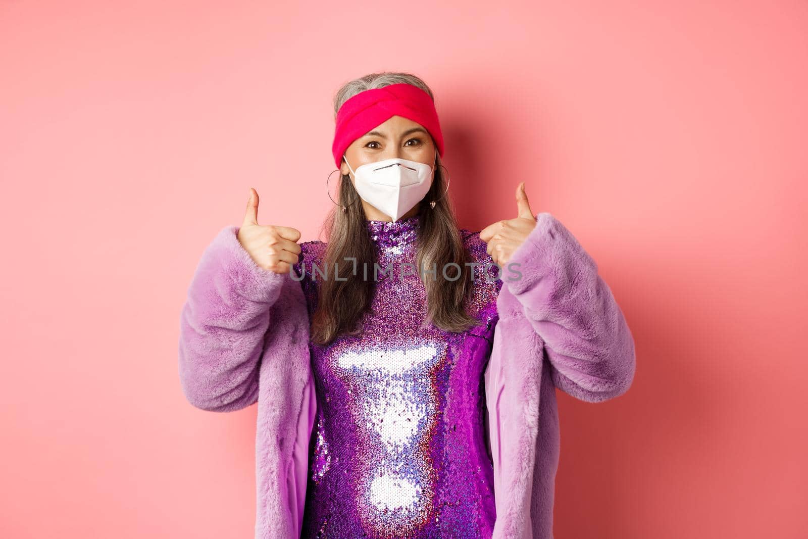 Covid-19, pandemic and fashion concept. Cheerful asian senior woman partying during coronavirus with respirator, showing thumbs-up, recommend wear face masks, pink background.