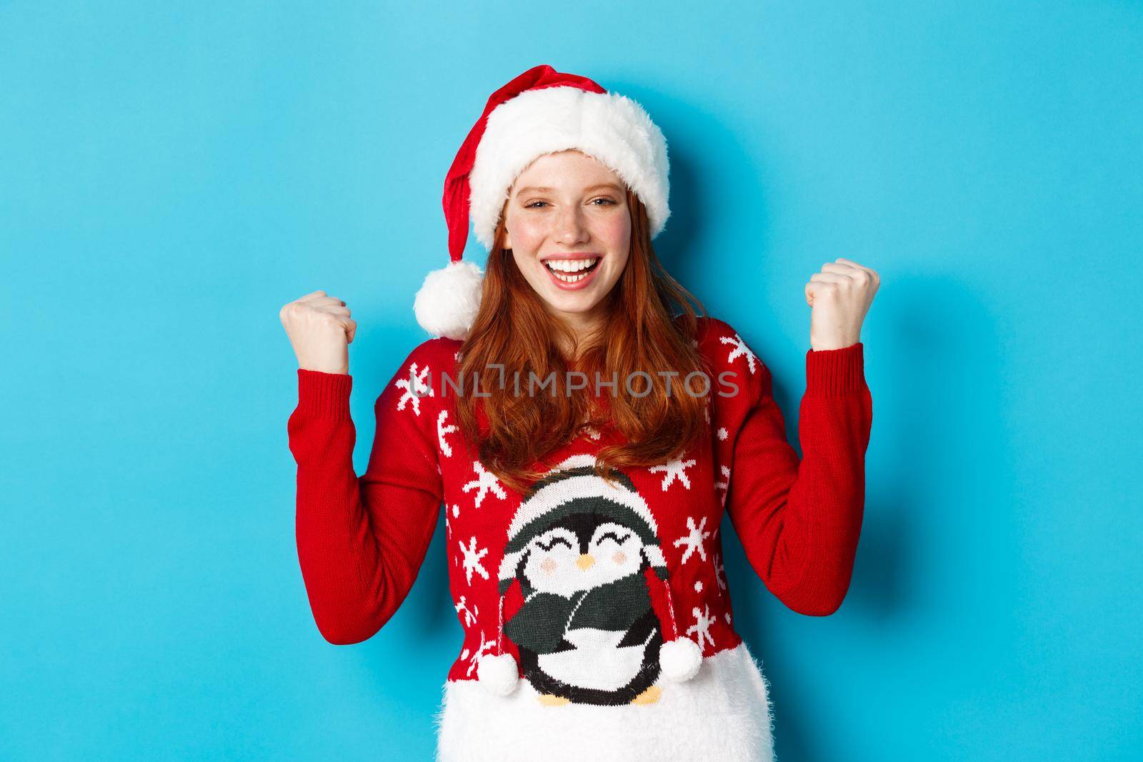 Happy holidays and Christmas concept. Cheerful redhead girl in santa hat and xmas sweater, winning and celebrating victory, raising hands up satisfied, triumphing by Benzoix