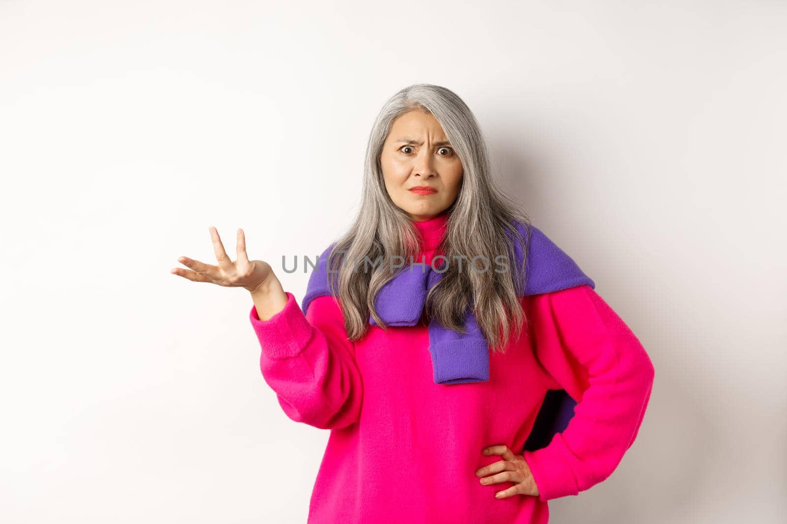 Angry and confused asian senior woman spread hand sideways and staring at camera puzzled, standing in pink sweater against white background by Benzoix