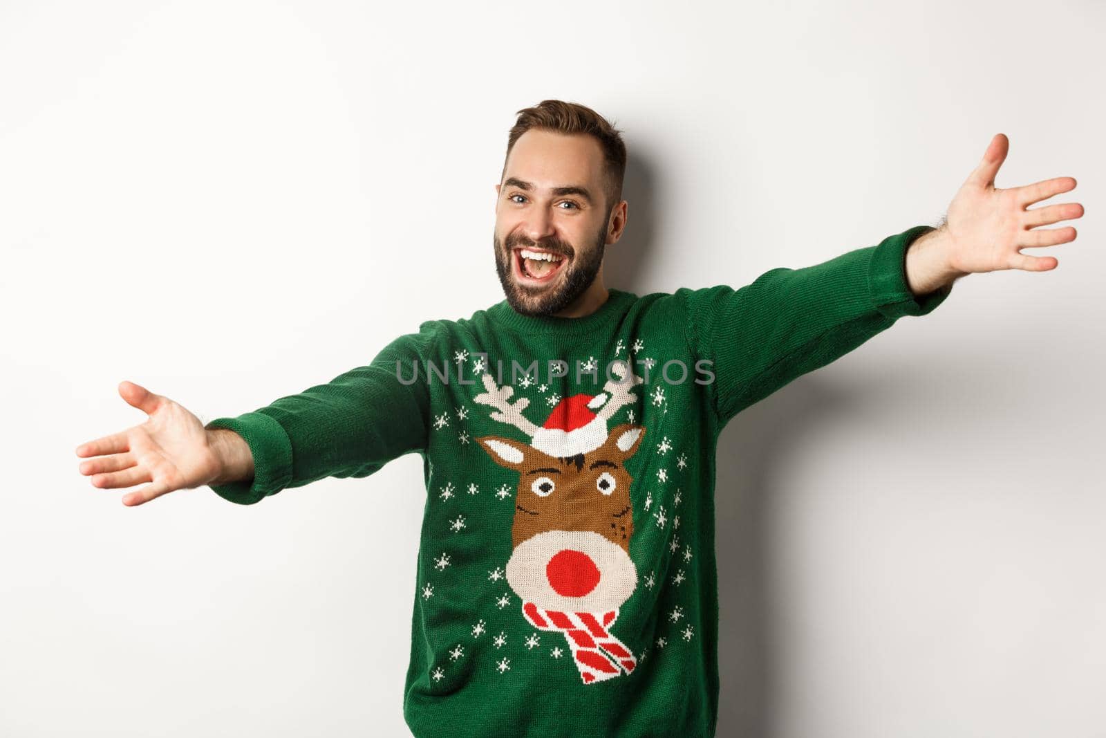 New year celebration and winter holidays concept. Friendly man spread hands for hug, welcome to Christmas party, wearing funny sweater, white background.