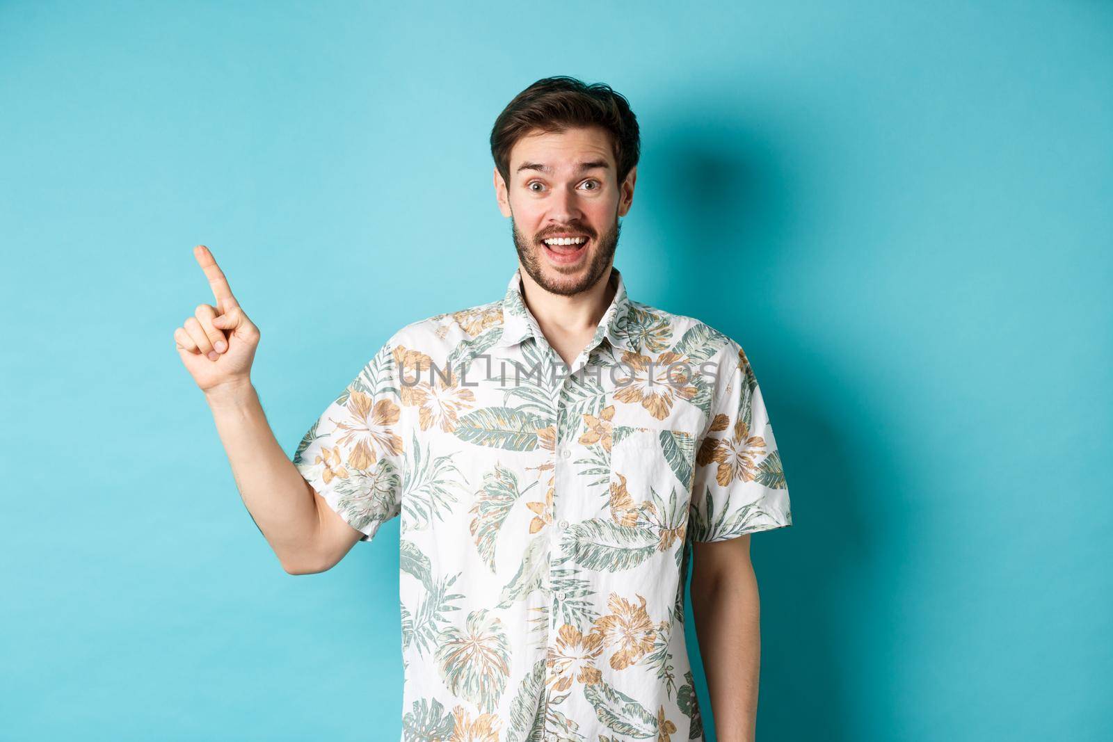Surprised happy tourist pointing finger left at empty space, showing cool promo, standing in hawaiian shirt on blue background.