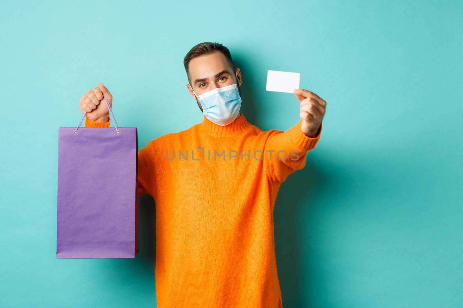 Covid-19, pandemic and lifestyle concept. Happy male customer in face mask showing credit card and purple shopping bag, standing over light blue background by Benzoix