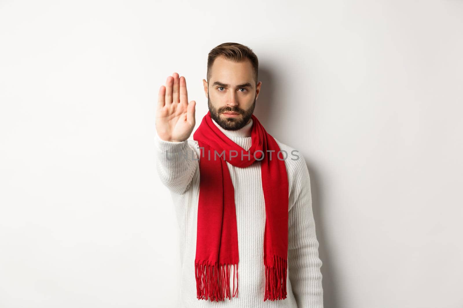 Serious bearded guy saying no, showing stop sign, rejection sign, prohibit action, standing in winter sweater and red scarf against white background.