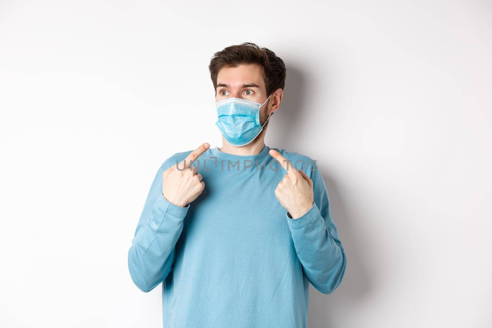 Coronavirus, health and quarantine concept. Confused young man pointing at medical mask on face and looking left, standing over white background.