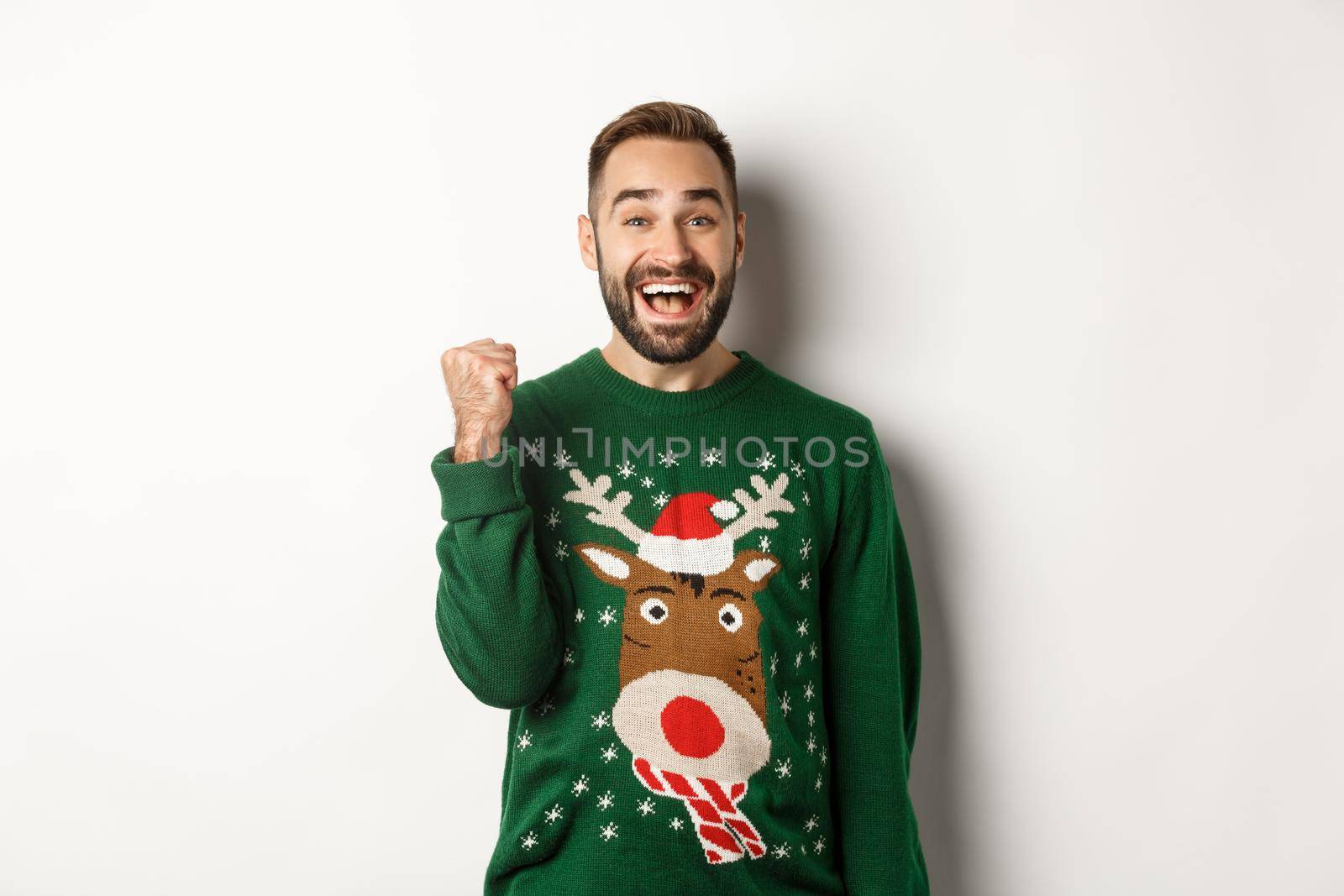 New Year, holidays and celebration. Excited and happy guy in Christmas sweater making fist pump, triumphing, receiving a gift and rejoicing, standing over white background by Benzoix