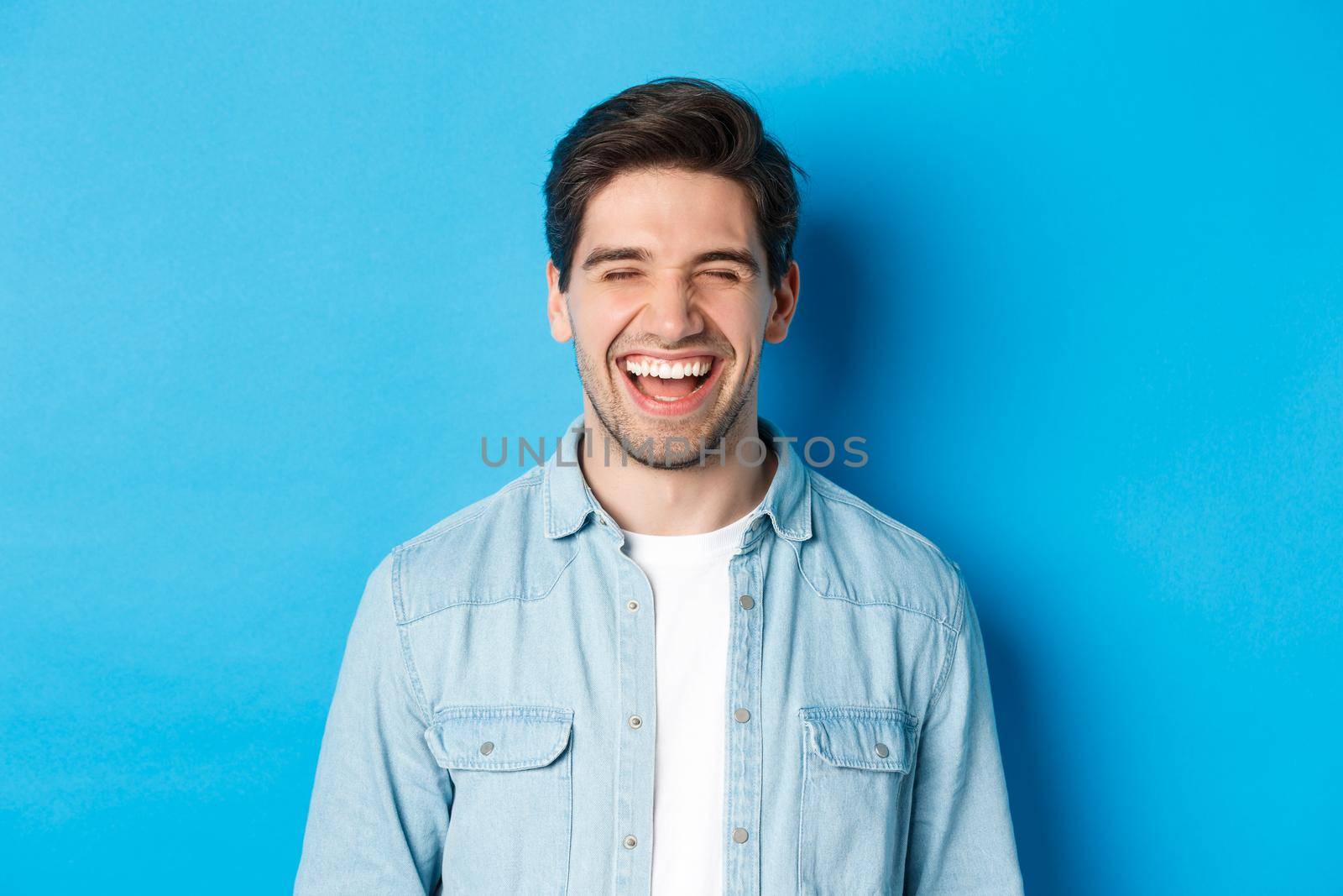 Close-up of happy young bearded man in casual clothes, laughing and feeling joyful, standing over blue background.
