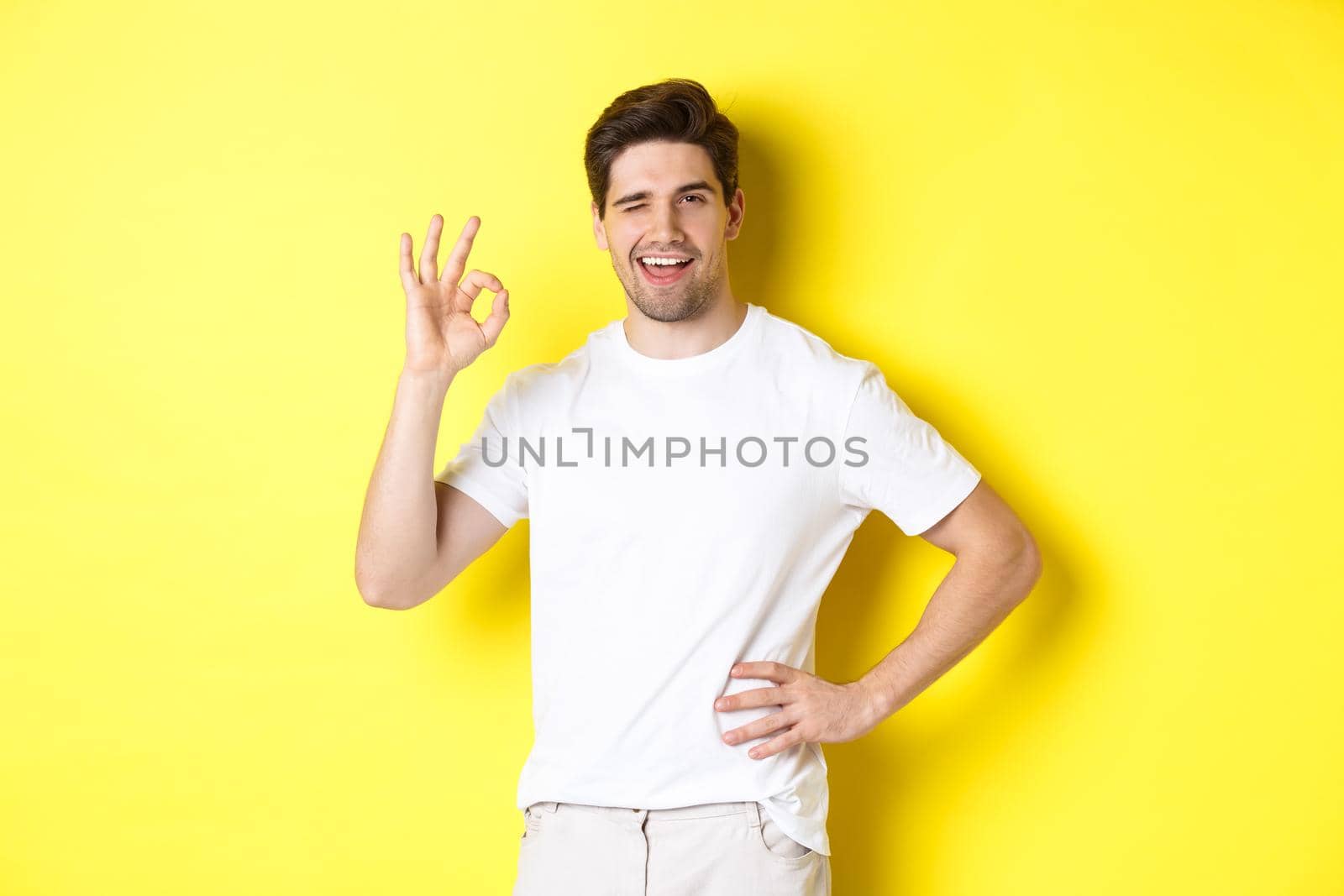 Confident handsome man winking, showing okay sign in approval, like something good, standing over yellow background by Benzoix
